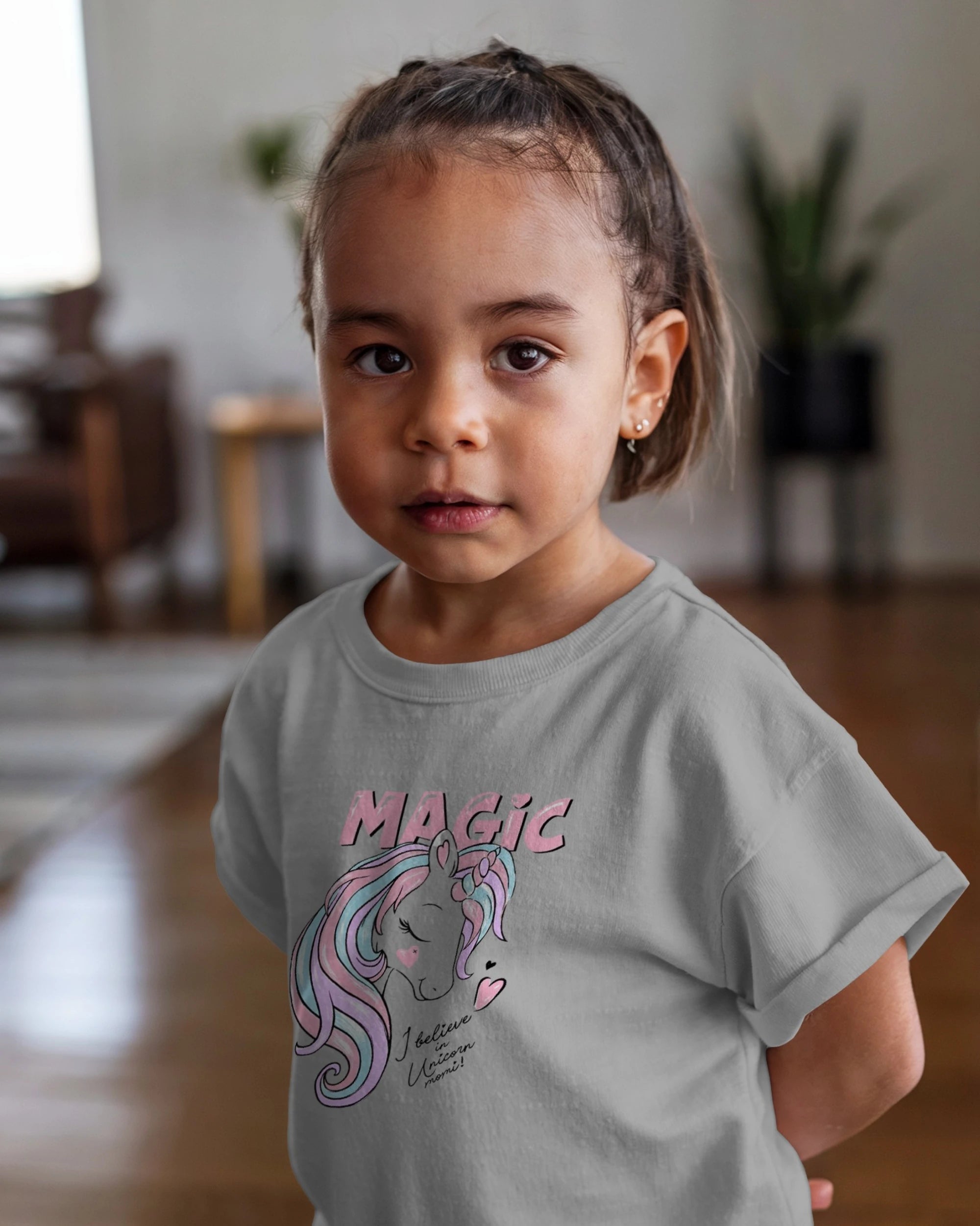 Child wearing a gray t-shirt with a unicorn graphic and 'Magic' text in a home setting.