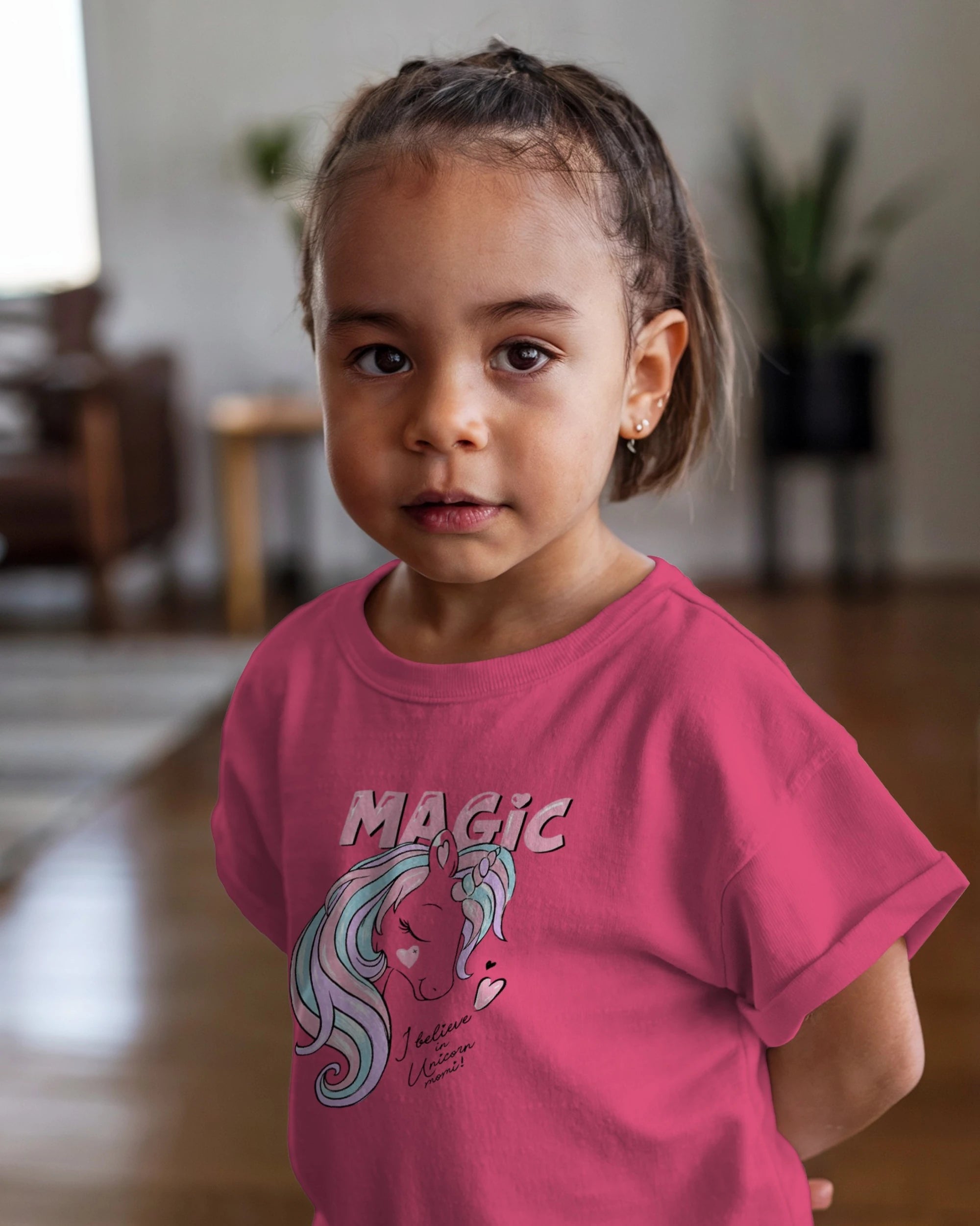 Young girl wearing a pink t-shirt with a unicorn graphic and 'MAGIC' text indoors.