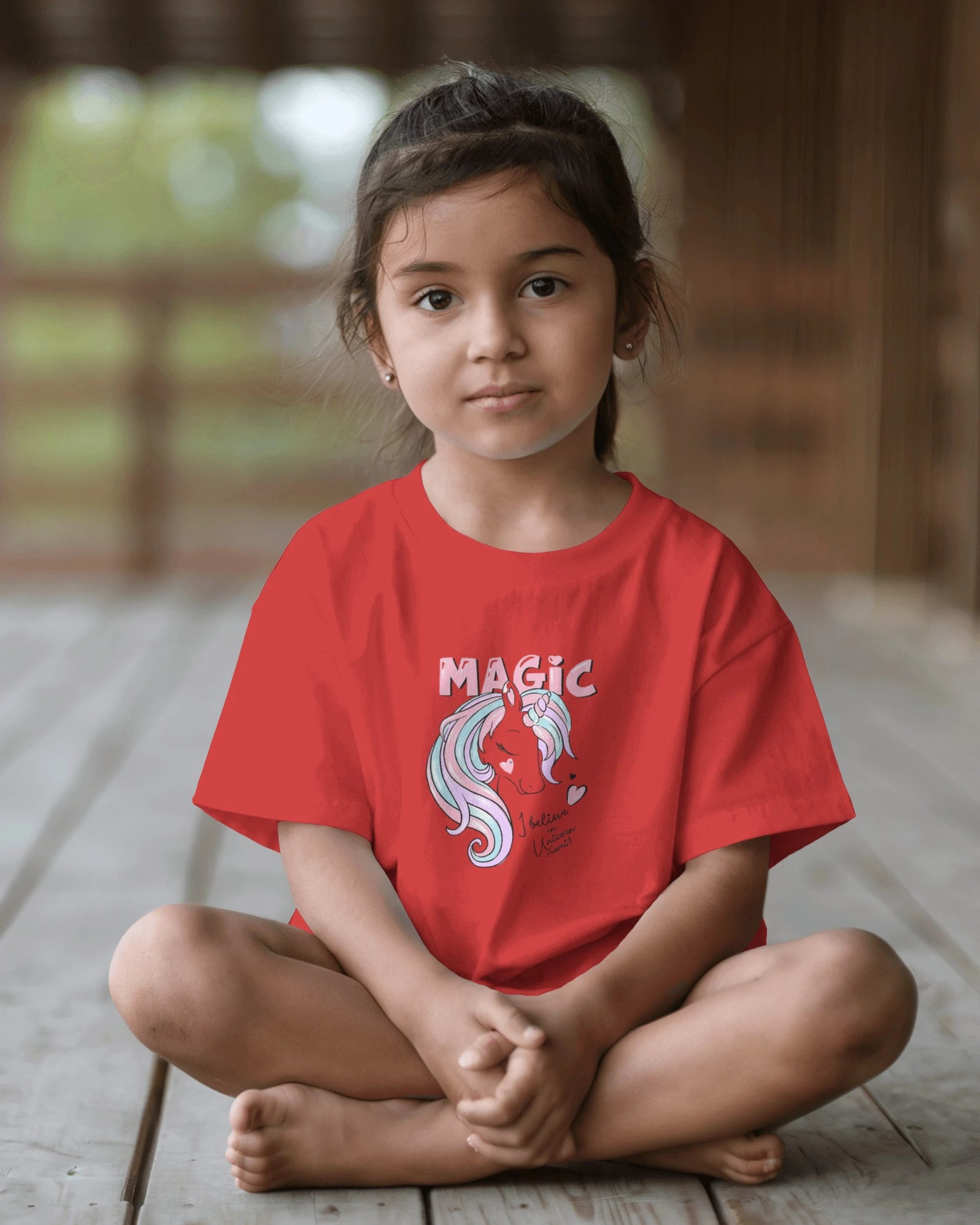 Child wearing a red t-shirt with 'MAGIC' and a unicorn design, sitting on a wooden floor.
