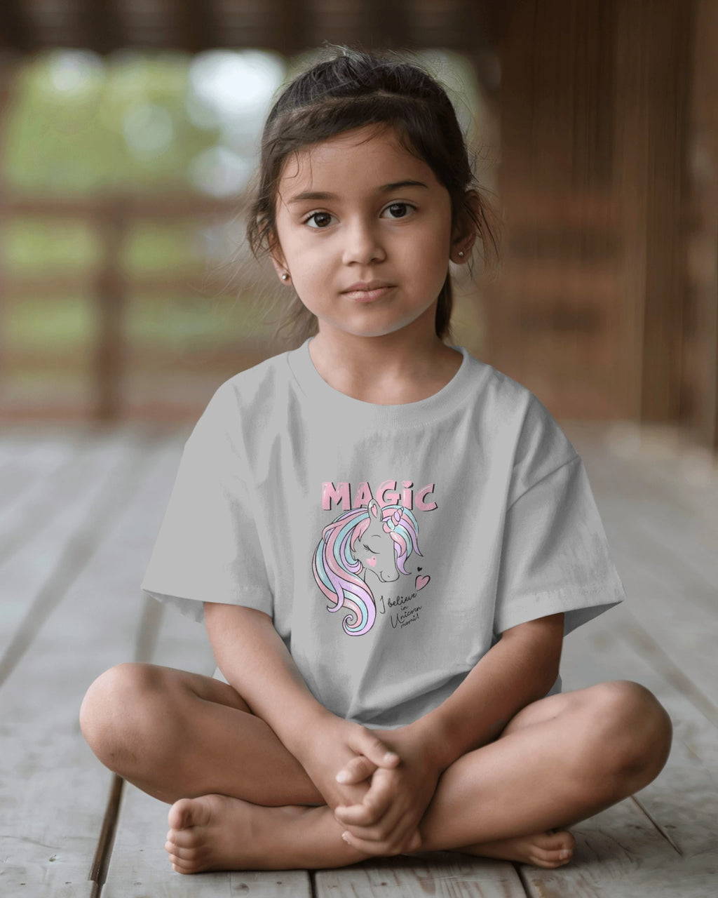 Young girl sitting on a wooden floor wearing a gray t-shirt with a unicorn design and 'Magic' text.