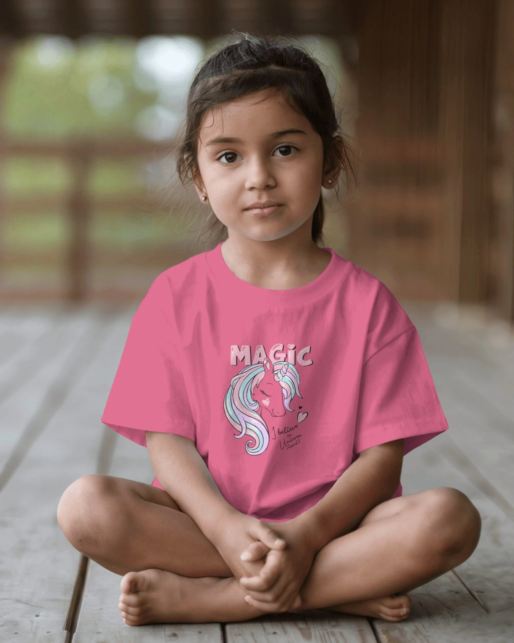 Young girl wearing a pink t-shirt with a unicorn graphic and 'MAGIC' text, sitting on a wooden floor.