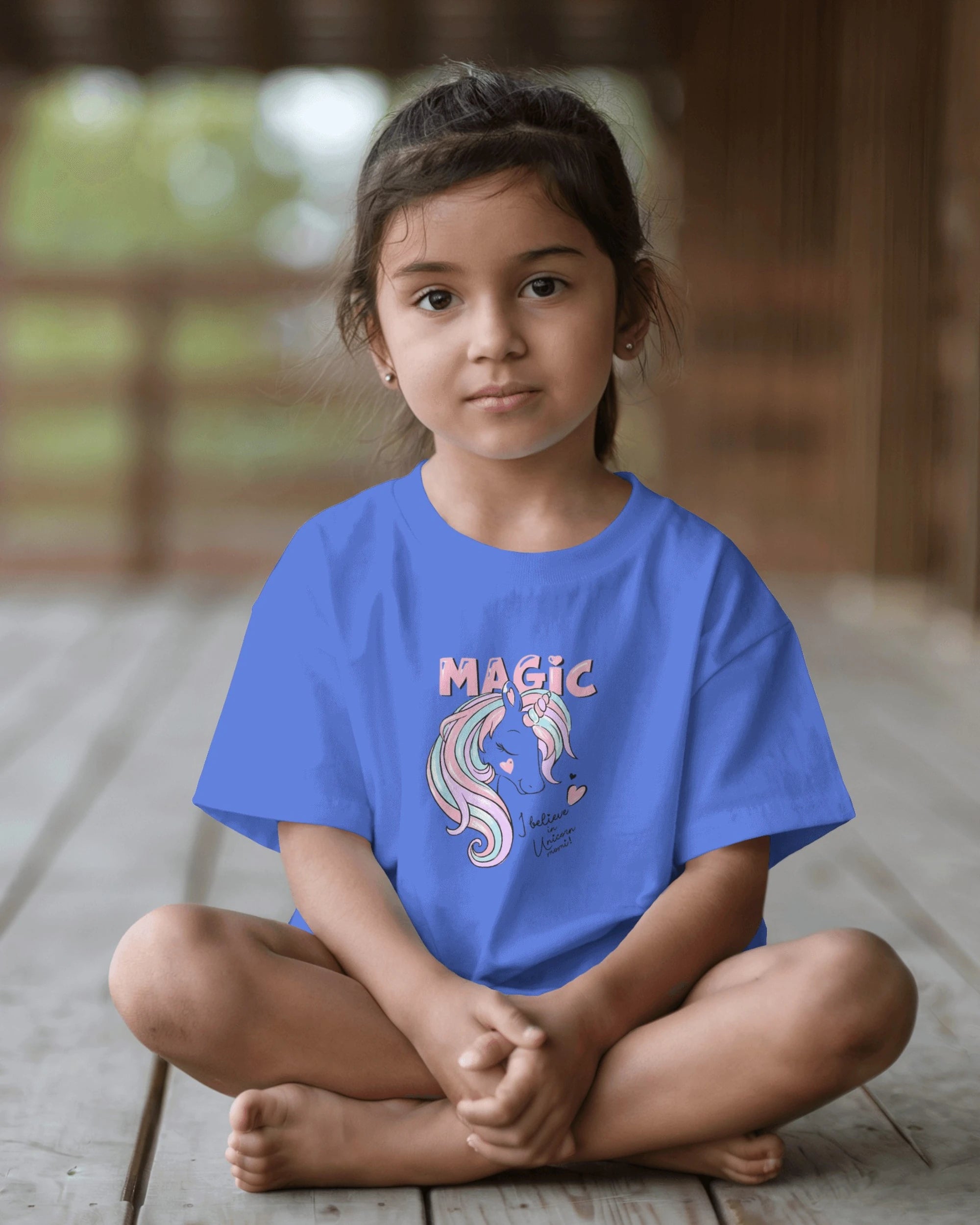 Child wearing a blue t-shirt with a unicorn graphic and 'MAGIC' text, sitting on a wooden floor.