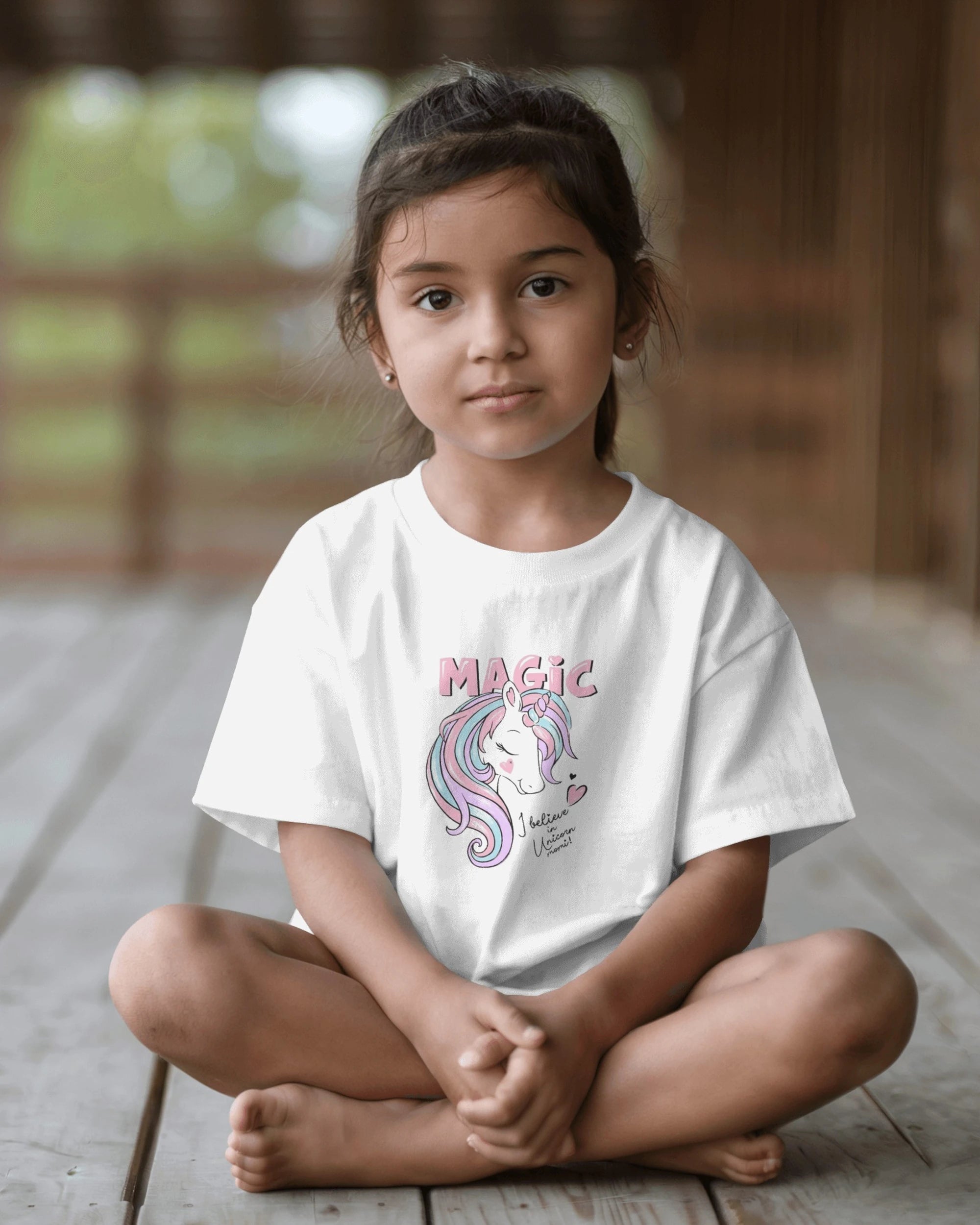 Young girl sitting on a wooden floor wearing a white t-shirt with a unicorn design and 'Magic' text.