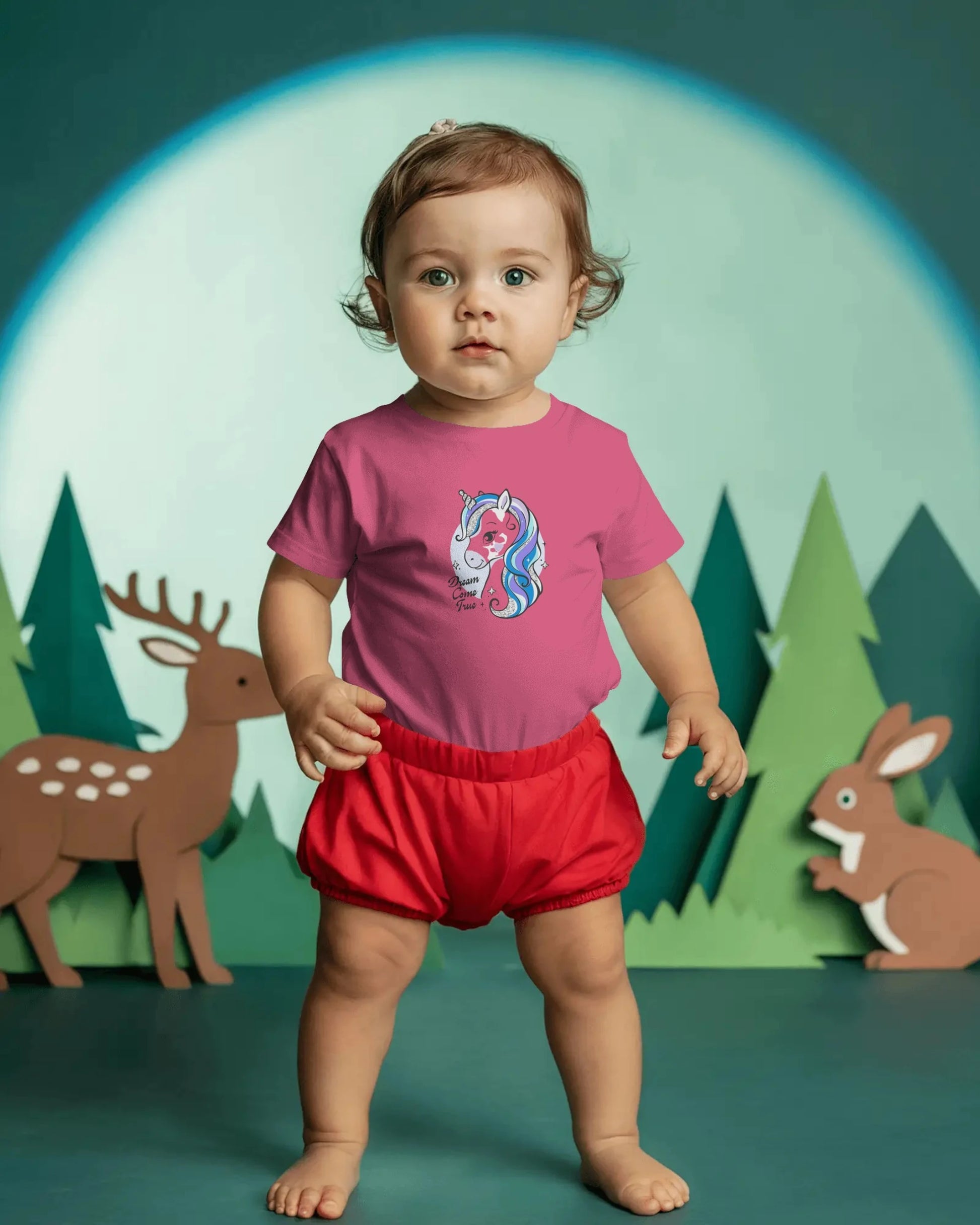 Child wearing a pink shirt with a unicorn design and red shorts in front of a forest background.