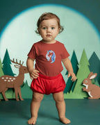 Child wearing a red shirt with a unicorn design and red shorts in front of a forest background