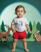 Child wearing a white shirt with a blue design and red shorts in front of a forest background