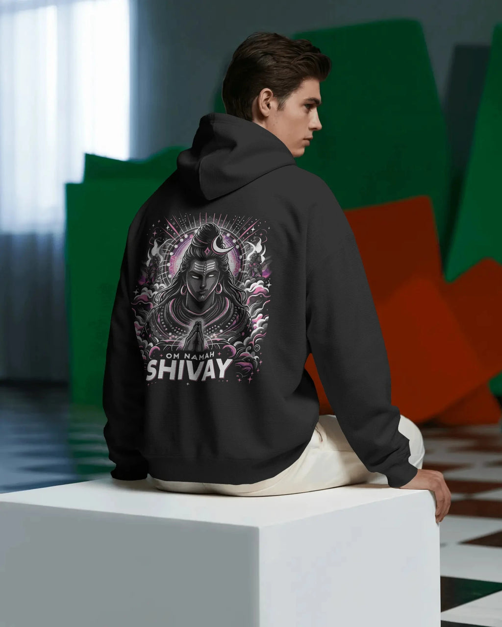 Person wearing a black pullover hoodie with a graphic design and text on the back, sitting on a white cube.