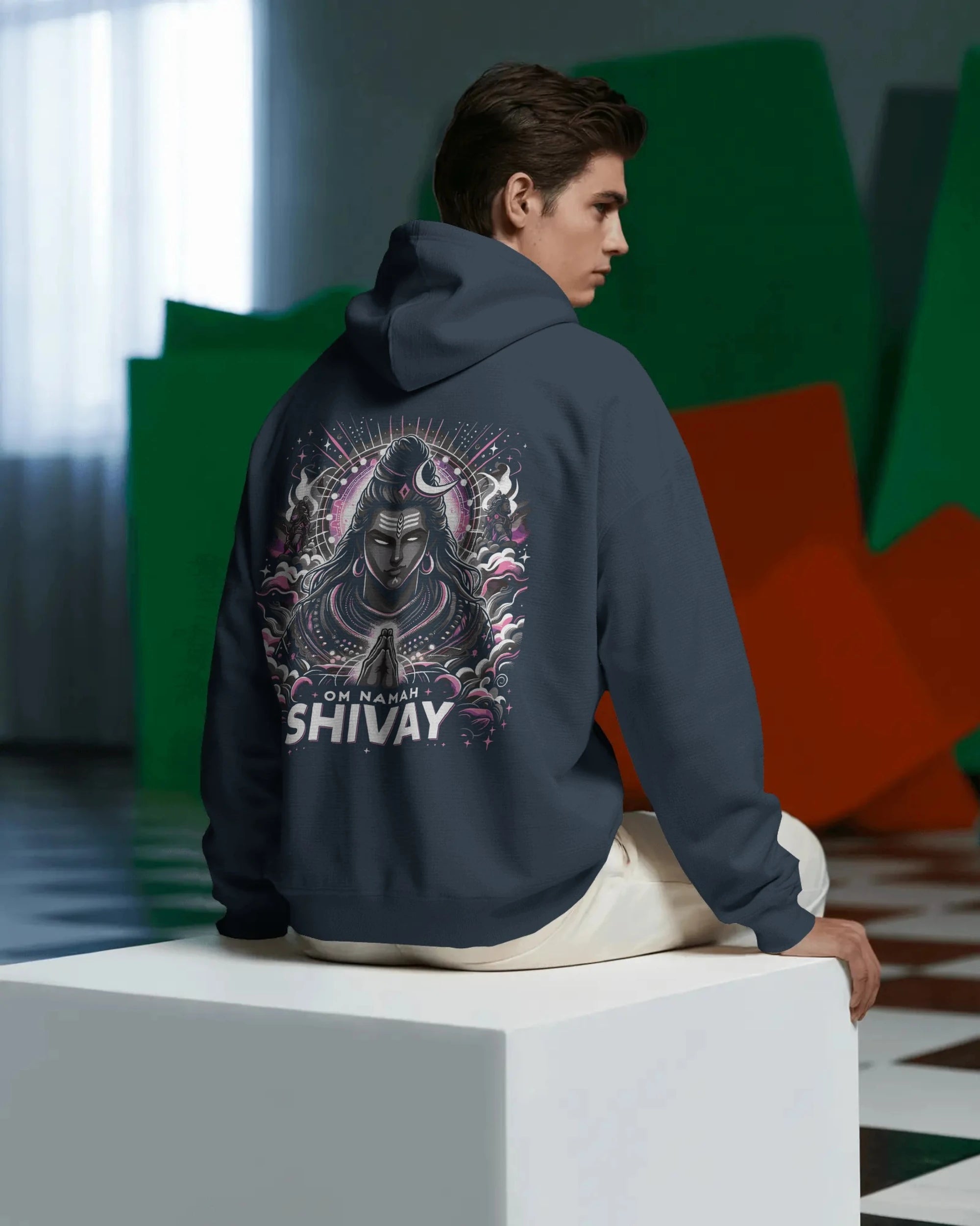 Person wearing a pullover hoodie with a 'Shivay' design on the back, sitting on a white block.