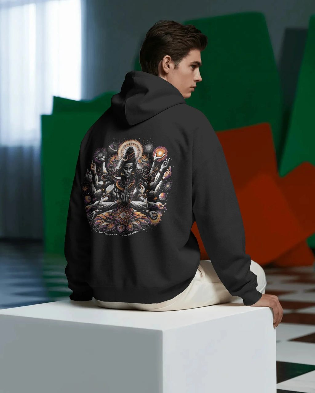 Person wearing a black pull over hoodie with a colorful design on the back, sitting on a white cube.