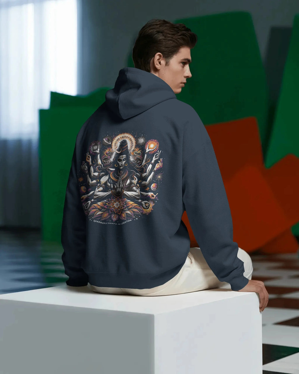 Person wearing a pull over  hoodie with a colorful design on the back, sitting on a white block.